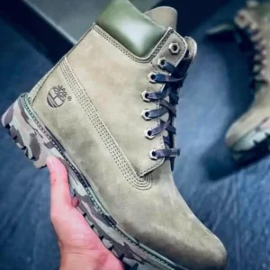Bottine montante beige avec col vert et semelle camouflage, tenue à la main sur un fond sombre.