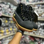 Bottines noires montantes avec semelle crantée marron et œillets dorés, tenues à la main dans un magasin.