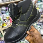 Chaussure de sécurité noire montante avec coutures jaunes, tenue à la main dans un magasin de chaussures.