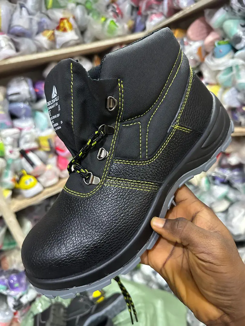 Chaussure de sécurité noire montante avec coutures jaunes, tenue à la main dans un magasin de chaussures.