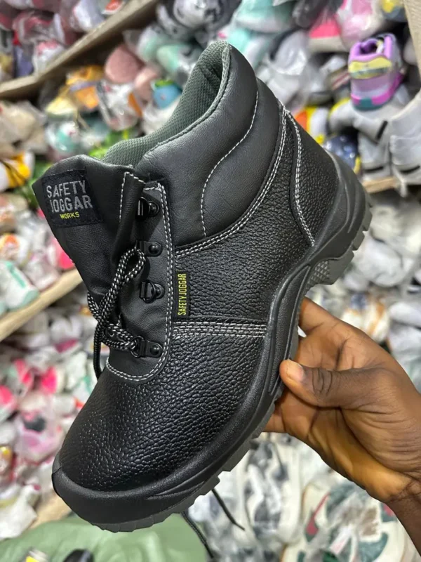Chaussure de sécurité noire montante – maintien & résistance Chaussure de sécurité noire à tige montante et lacets, tenue à la main dans un magasin, fond rempli de chaussures emballées.