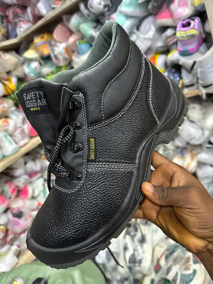 Chaussure de sécurité noire à tige montante et lacets, tenue à la main dans un magasin, fond rempli de chaussures emballées.