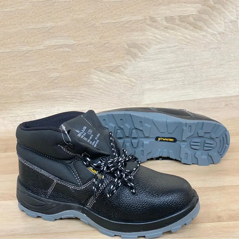 Chaussures de sécurité montantes noires à lacets, semelle grise crantée, posées sur un sol en bois.